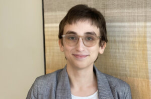 Professional headshot of Kayle Hill. A person with short brown hair and light skin, wearing clear-framed glasses and a gray plaid blazer over a white shirt, smiling softly while standing in front of a textured, neutral-toned background.