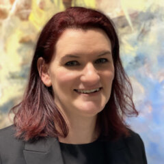 http://Headshot%20of%20Jacqueline%20Cadman.%20She%20has%20shoulder-length%20dark%20red%20hair%20and%20is%20wearing%20a%20black%20blazer%20over%20a%20black%20top.%20She%20is%20smiling%20and%20standing%20in%20front%20of%20a%20softly%20blurred,%20colorful%20abstract%20background.