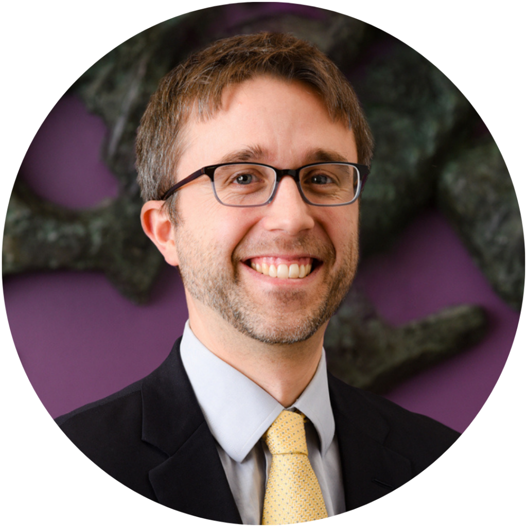Professional headshot of partner Jamie Strawbridge. A headshot of a smiling man wearing glasses, a dark suit, a light gray shirt, and a yellow tie. He has short brown hair and a trimmed beard. The background is a blurred image of dark grey abstract shapes against a purple wall.