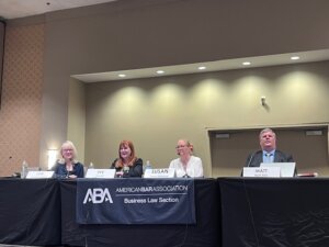 Four panelists are seated behind a table at an American Bar Association (ABA) Business Law Section event. Each has a nameplate in front of them: from left to right, the panelists are identified as Lori Sommerfield, Eve Hill, Susan Desmond, and Matt Ater. The backdrop is a plain beige wall with overhead lighting. A banner on the table reads "ABA American Bar Association Business Law Section." The panelists appear engaged and are using microphones.