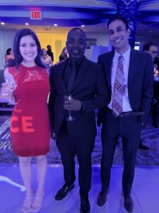 Three people dressed in formal attire are smiling and posing for a photo at an indoor event with dim blue lighting. The woman on the left is wearing a red dress and holding a wine glass. The man in the center is wearing an all-black suit and also holding a wine glass. The man on the right is wearing a dark suit with a white shirt and plaid tie. Other guests and event decor are visible in the background.
