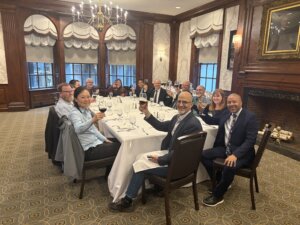 A group of thirteen people are seated around a long, white-clothed dining table in an elegant wood-paneled room with large windows and a chandelier. They are smiling and raising glasses in a toast during a formal dinner. The atmosphere is warm and collegial, reflecting a celebratory gathering. This image was taken during the “Born-Accessible Design for Digital Accessibility” convening workshop at the Harvard Radcliffe Institute on May 5–6.