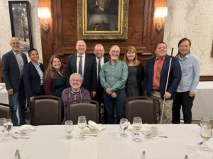 Ten individuals are standing at the front of a formal dining room, smiling for a group photo under a framed portrait and flanked by wood-paneled walls. The group includes leaders in disability and technology who participated in the “Born-Accessible Design for Digital Accessibility” workshop at the Harvard Radcliffe Institute in Boston on May 5–6.