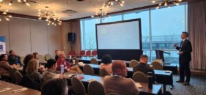 Partner Jamie Strawbridge presents to a group of seated attendees in a modern conference room with large windows. The audience is attentively listening, and a projection screen displays a presentation titled "Document Accessibility: A Legal Imperative, Staying Compliant in 2025." The room features mid-century modern light fixtures and an outdoor view.