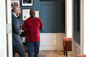 Alt text: Two men, Governor Wes Moore and exoneree Tyrone Jones, stand conversing and laughing in a warmly lit office space with blue walls and white paneling. Governor Wes Moore wears a gray suit, and Tyrone Jones wears a red polo shirt and jeans. Behind them are framed certificates, photographs, and a bookshelf.