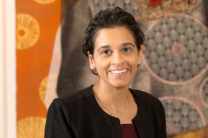 Headshot for new Brown, Goldstein & Levy attorney Aranda Stathers. She has short black hair and smiles warmly at the camera. She is wearing a black blazer over a burgundy shirt in front of an orange and gray abstract background.