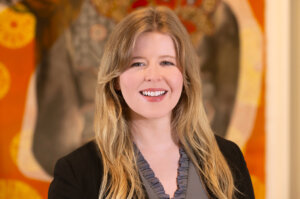 Headshot for new Brown, Goldstein & Levy attorney Samantha Westrum. She has long blonde hair and smiles warmly at the camera. She is wearing a black blazer over a ruffled gray blouse in front of an orange abstract background.