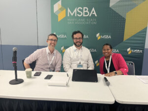 Three BGL attorneys - Kevin Docherty, Anisha Queen, and Jamie Strawbridge - smile while seated at a long table in front of a Maryland State Bar Association (MSBA) banner during the 2025 MSBA Legal Summit, where they presented a seminar. The table holds microphones, a coffee cup, phones, notepad, and computer.