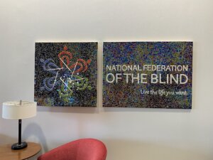 A wall-mounted mosaic art piece composed of two panels. The left panel features the logo for the National Federation of the Blind. The right panel displays the text "NATIONAL FEDERATION OF THE BLIND" and the tagline "Live the life you want." in white letters against a multicolored mosaic background. Below the panels are a red chair, a wooden side table, and a black lamp with a white lampshade.
