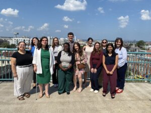 A group of Brown, Goldstein & Levy attorneys and staff members along with a few National Federation of the Blind team members stand on the patio rooftop of the National Federation of the Blind headquarters in Baltimore, MD. Behind the group is the Baltimore skyline with a blue sky and some white clouds. Everyone is smiling toward the camera and wearing business casual clothing.
