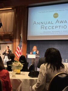 Eve Hill at a podium, gesturing with her hands while speaking to a large group. The backs of some audience members seated at tables can be seen seated in the foreground.