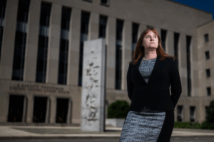 Eve Hill in the foreground, looking past the camera, standing in front of a government building.