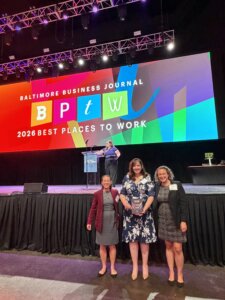 Three Brown, Goldstein & Levy team members (from left to right: Monica Basche, Rhonda Wedel, and Jessie Weber) pose with a "Best Places to Work" award in front of a stage at the Baltimore Business Journal's 2026 Best Places to Work ceremony. Behind them, a large, colorful screen displays "Baltimore Business Journal 2026 Best Places to Work," while a speaker stands at the podium on stage.